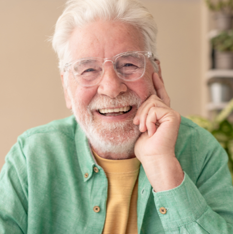 Friendly older man with white hair and beard, wearing glasses and a green shirt, smiling at the camera indoors.