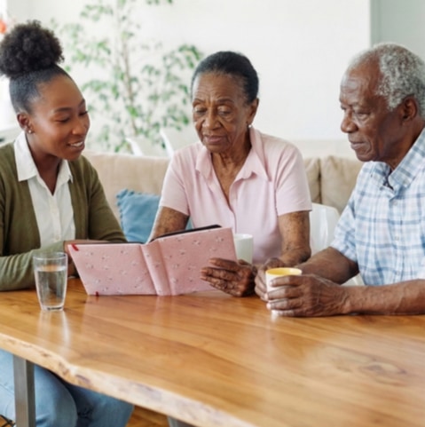 Young woman demonstrates a pink tablet to an elderly couple at a wooden dining table.