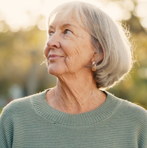 Older woman with gray hair looking upward, wearing a green sweater, outdoors in warm sunlight.