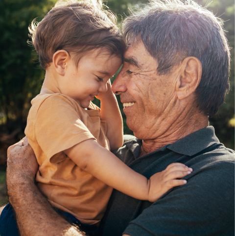 Grandfather holding a toddler close, both smiling and touching foreheads outdoors.