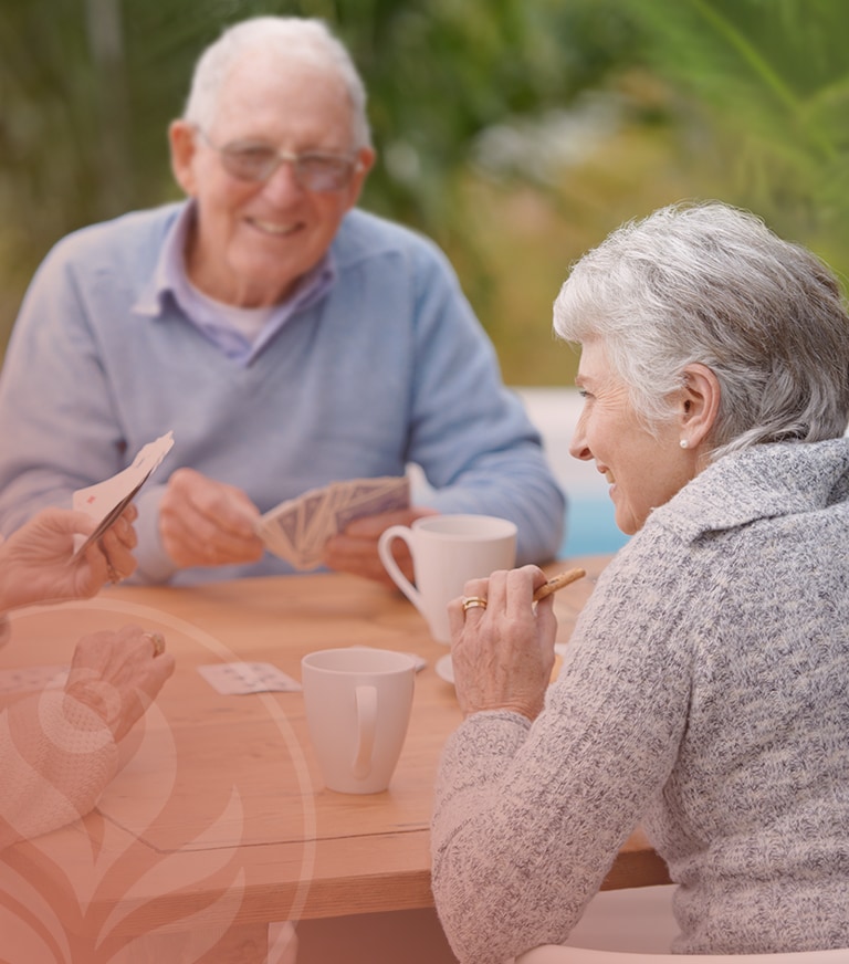 Two elderly people playing cards at a wooden outdoor table, smiling with mugs nearby on a sunny patio.