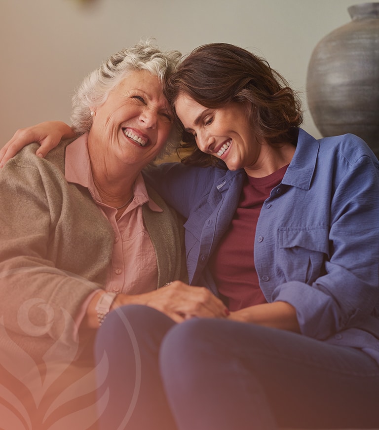 Two women sit close together, smiling and leaning their heads together in a warm, affectionate moment.