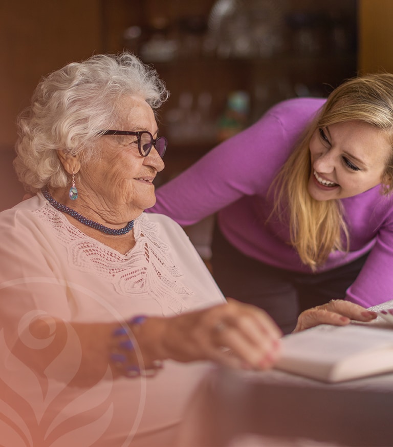 Older woman with white hair and glasses smiles as a younger caregiver leans in, helping with a book at a table in a home setting.