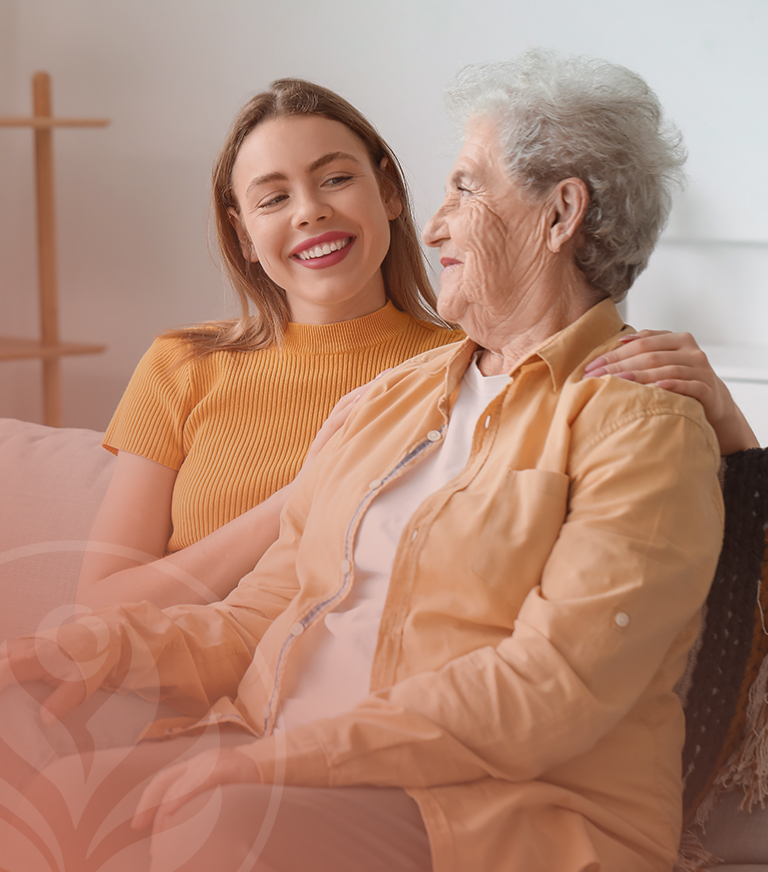 Two women, a younger and an older woman, sit close on a sofa smiling warmly at each other, showing companionship.