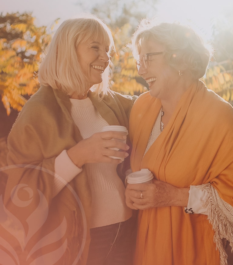 Two smiling older women enjoy coffee together outdoors on a sunny autumn day, sharing a warm moment of friendship.
