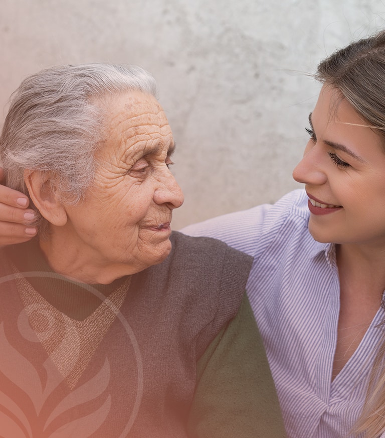 Young woman smiles at and comforts an elderly woman, gently leaning in for a warm, supportive moment.