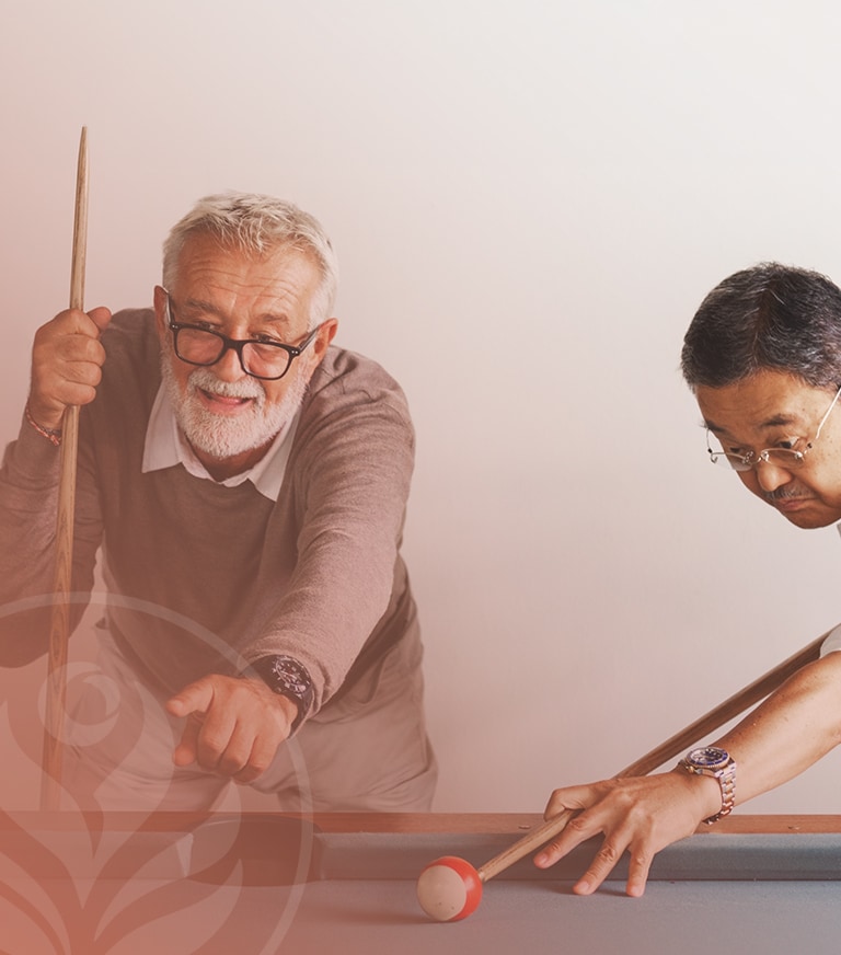 Two men play pool at a table; the man on the left with glasses points toward the cue ball while the other prepares a shot in the right foreground.