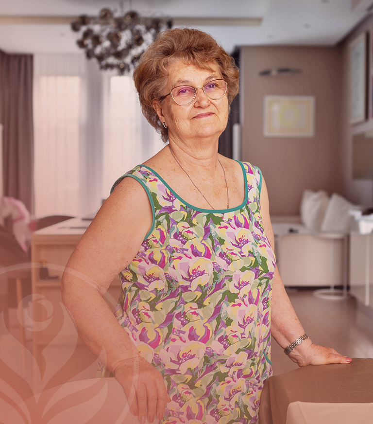 Older woman with short brown hair and glasses, wearing a colorful floral sleeveless dress, standing by a table in a bright living room.